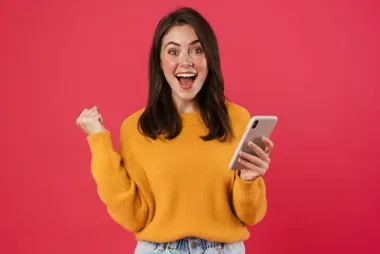 Uśmiechnięta kobieta w żółtym swetrze trzyma telefon i unosi zaciśniętą pięść na różowym tle.